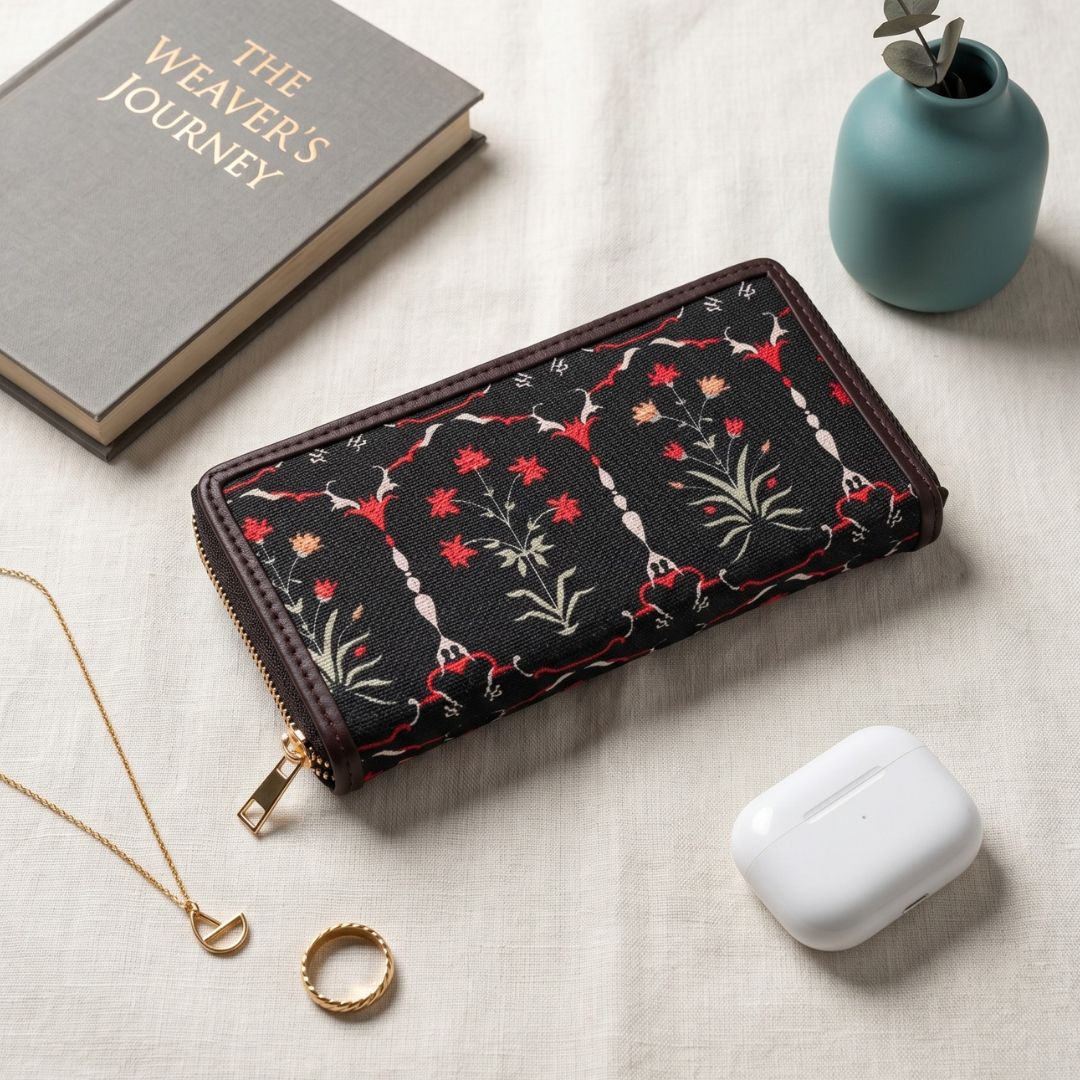 A black zip-around wallet with red, green, and white floral embroidery, styled on a beige linen surface with a gray book, a small teal vase with eucalyptus, a gold necklace, a gold ring, and a white wireless earbud charging case.