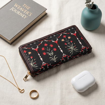 A black zip-around wallet with red, green, and white floral embroidery, styled on a beige linen surface with a gray book, a small teal vase with eucalyptus, a gold necklace, a gold ring, and a white wireless earbud charging case.